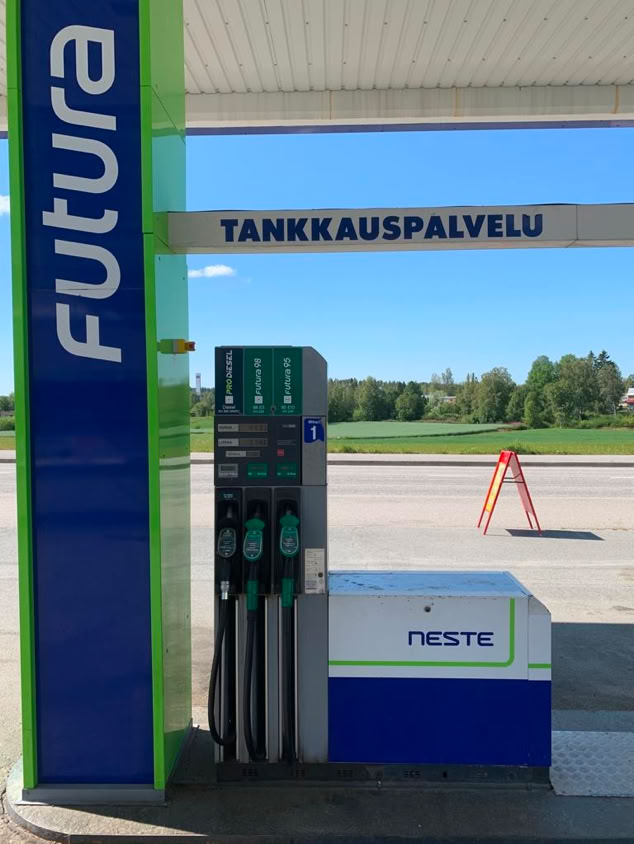 Gas station pump with "Futura" branding, displaying multiple fuel options, and a sign reading "TANKKAUSPALVELU." A green landscape is visible in the background.
