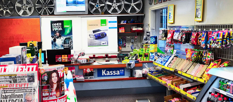 A convenience store checkout area with a counter labeled "Kassa," surrounded by shelves displaying various snacks, magazines, and small items.